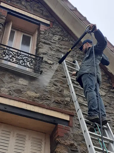 Couvreur de FM Couverture 77 nettoyant une façade en pierre et les abords de toiture à l’aide d’un nettoyeur haute pression depuis une échelle